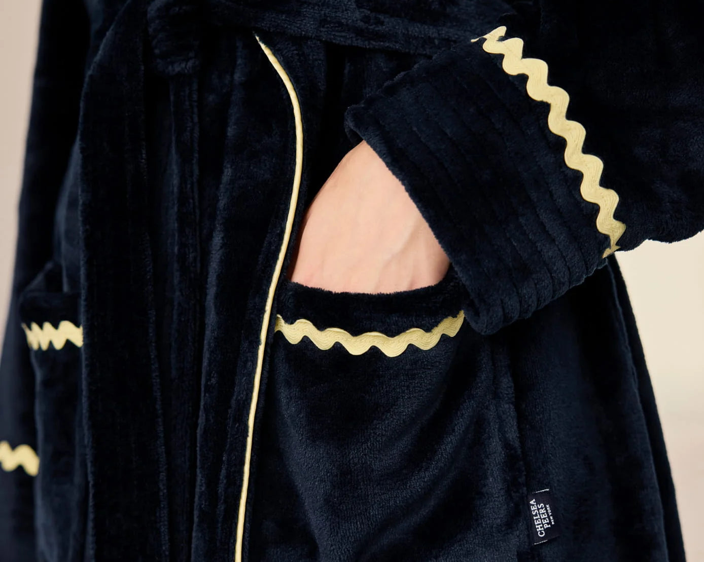 Close-up of a plush dark navy velvet robe with gold zigzag trim and a matching tie belt, shown on a model with one hand in the pocket.