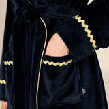 Close-up of a plush dark navy velvet robe with gold zigzag trim and a matching tie belt, shown on a model with one hand in the pocket.