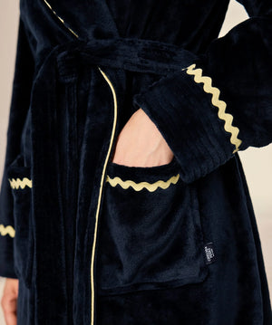 Close-up of a plush dark navy velvet robe with gold zigzag trim and a matching tie belt, shown on a model with one hand in the pocket.