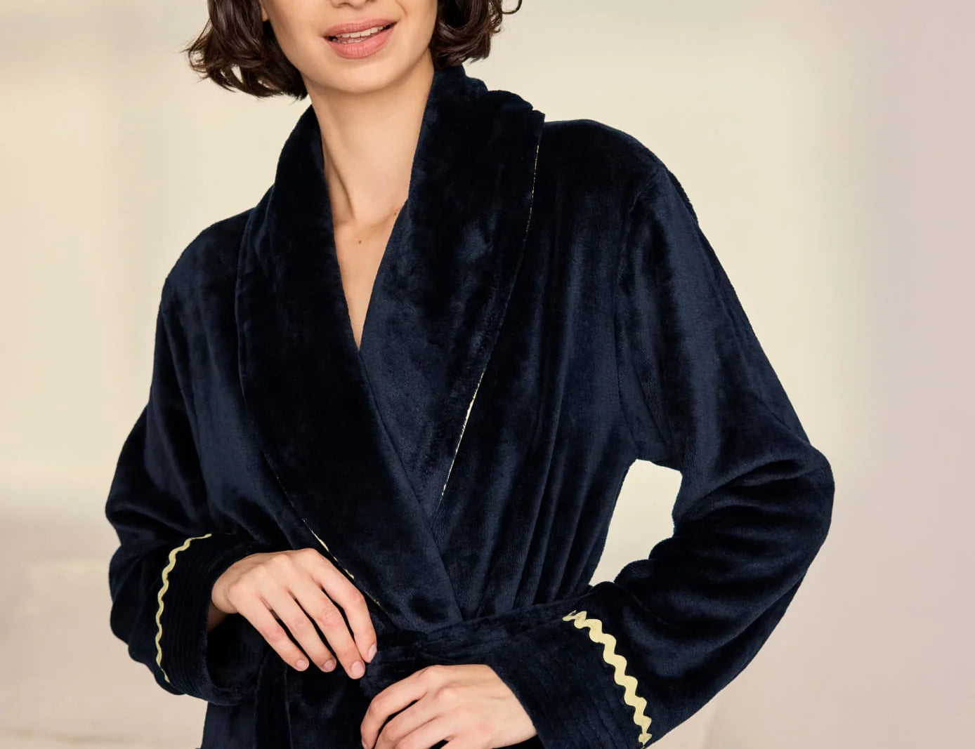 A woman wearing a plush navy blue velvet bathrobe with gold trim, standing against a soft beige background.