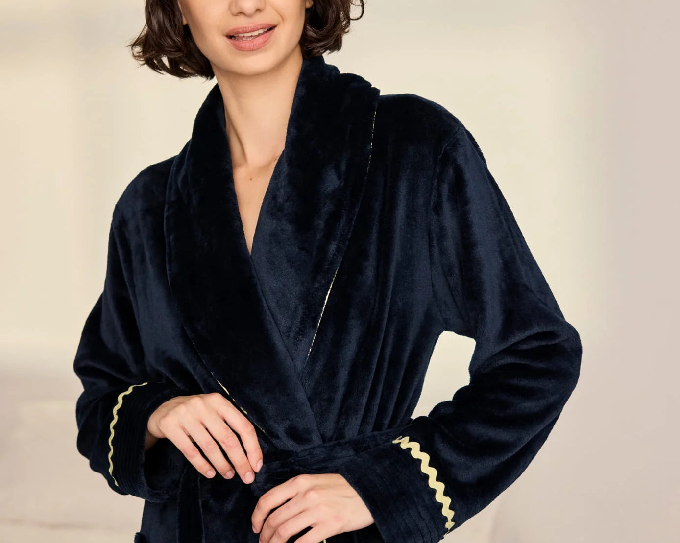 A woman wearing a plush navy blue velvet bathrobe with gold trim, standing against a soft beige background.