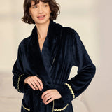 A woman wearing a plush navy blue velvet bathrobe with gold trim, standing against a soft beige background.