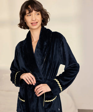 A woman wearing a plush navy blue velvet bathrobe with gold trim, standing against a soft beige background.