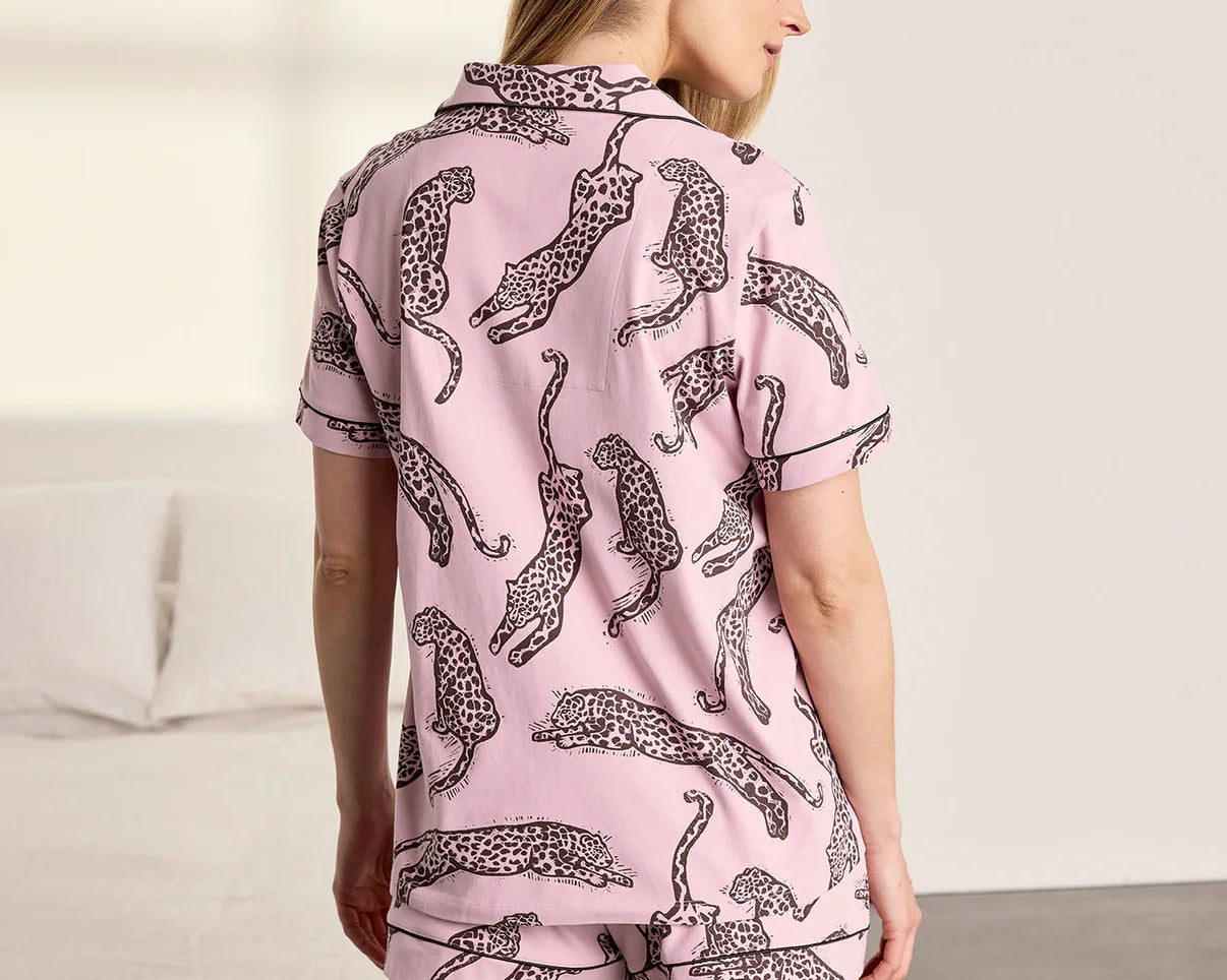 A woman wearing a light pink short-sleeve pajama set with a black leopard print, shown from the back in a bedroom setting.
