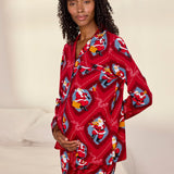A woman wearing a red long-sleeve pajama set with a festive Santa Claus print, standing in a bedroom setting with a bed in the background.