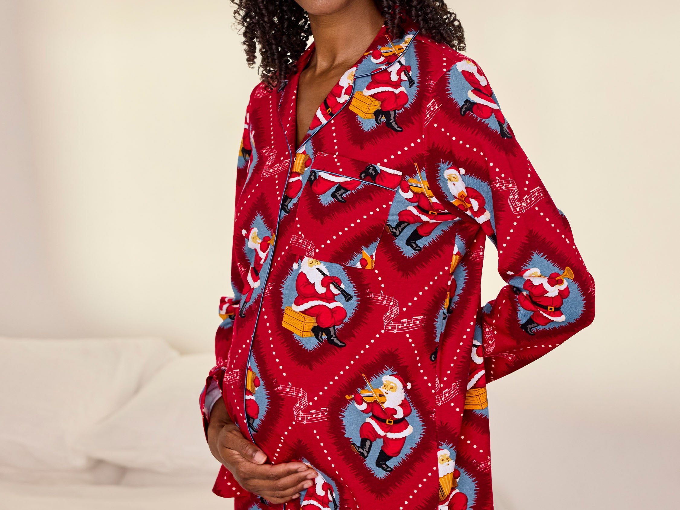 A woman wearing a red long-sleeve pajama set with a festive Santa Claus print, standing in a bedroom setting with a bed in the background.