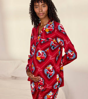 A woman wearing a red long-sleeve pajama set with a festive Santa Claus print, standing in a bedroom setting with a bed in the background.