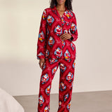 A woman stands barefoot in a studio wearing a red long-sleeve pajama set with an allover Santa Claus print. The set includes a button-front top and matching pants.
