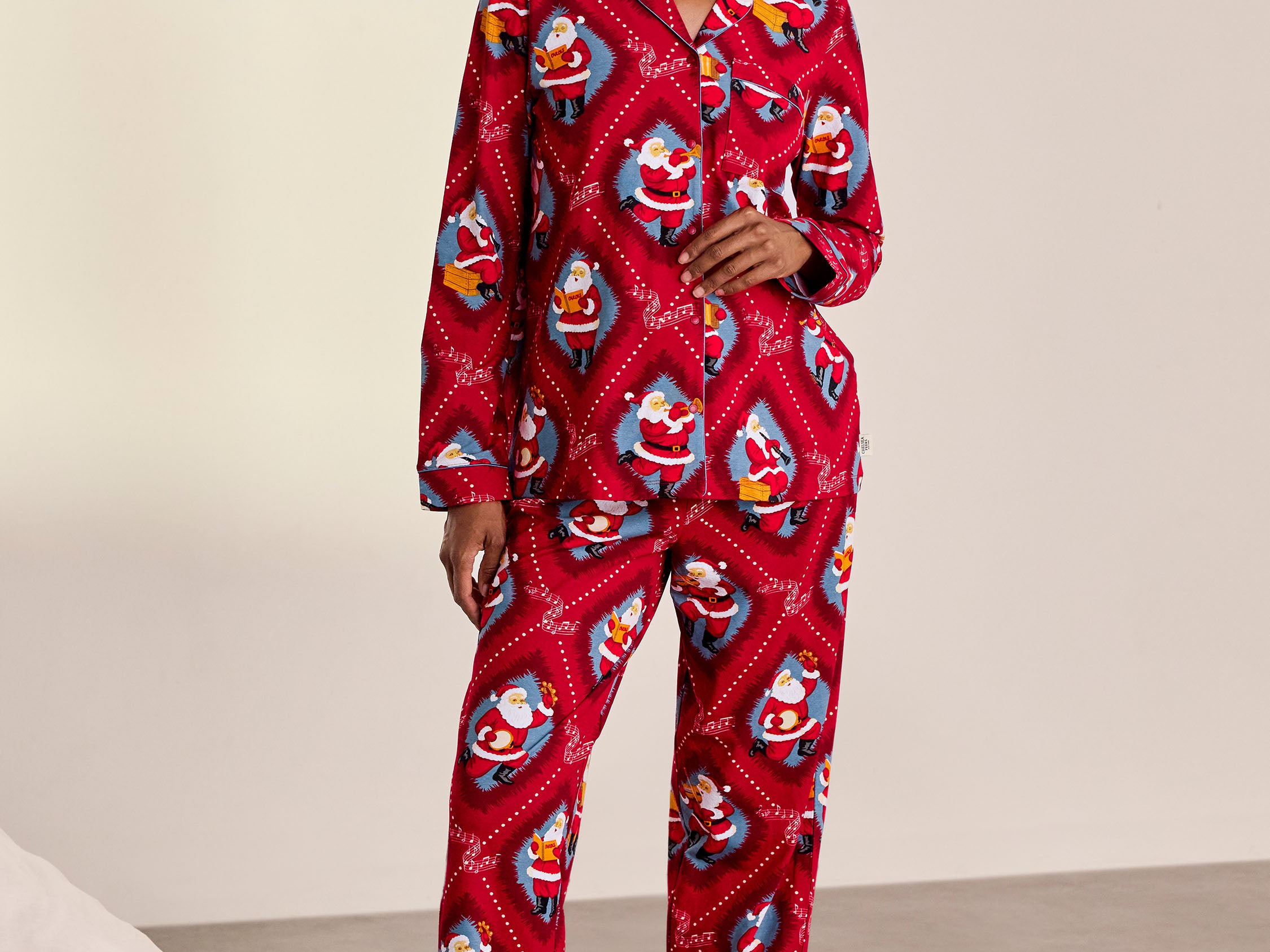 A woman stands barefoot in a studio wearing a red long-sleeve pajama set with an allover Santa Claus print. The set includes a button-front top and matching pants.