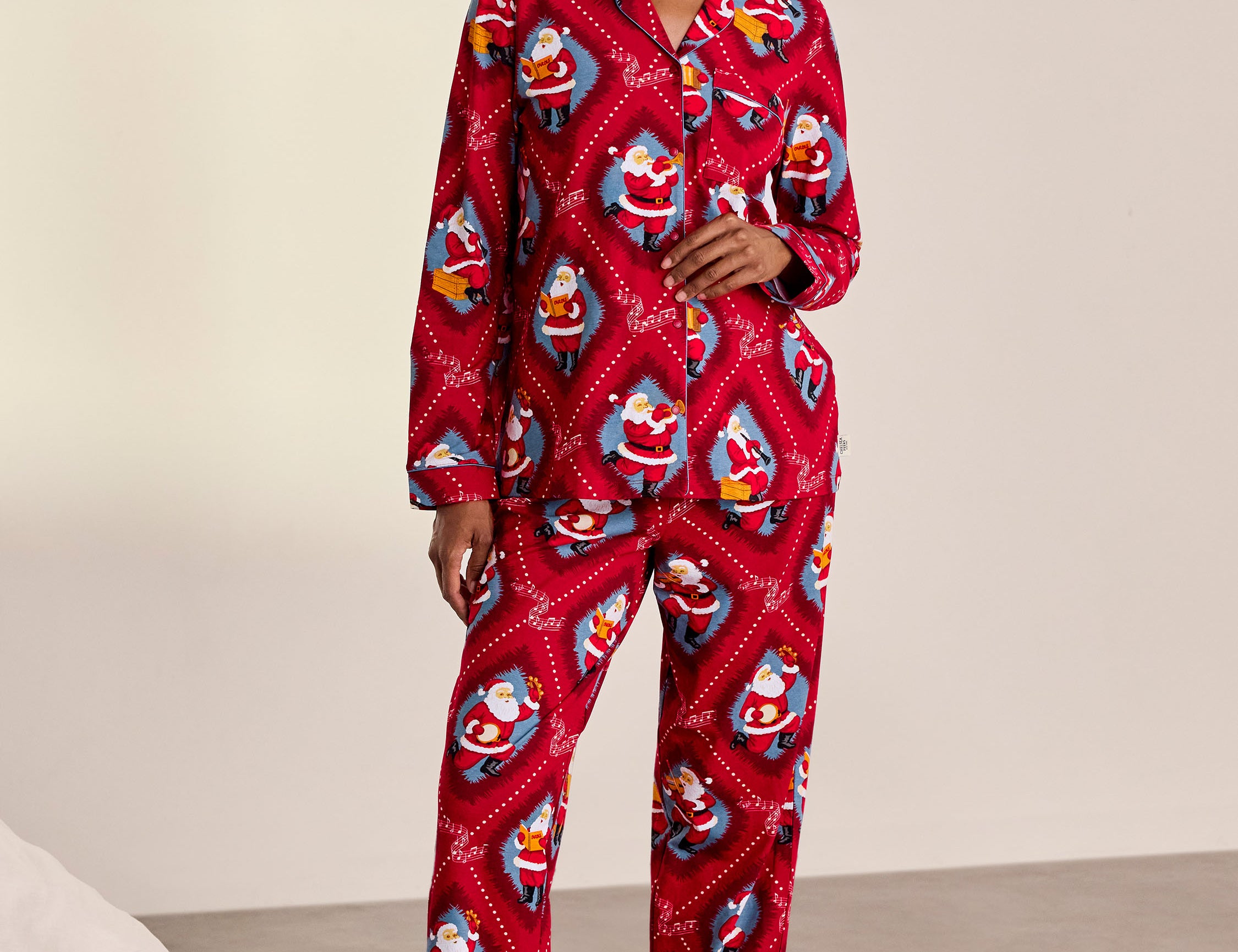 A woman stands barefoot in a studio wearing a red long-sleeve pajama set with an allover Santa Claus print. The set includes a button-front top and matching pants.