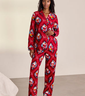 A woman stands barefoot in a studio wearing a red long-sleeve pajama set with an allover Santa Claus print. The set includes a button-front top and matching pants.