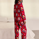 A woman stands barefoot in a bedroom wearing a red long-sleeve pajama one-piece covered in a repeating Santa Claus print. A white bed and light wall are visible in the background.