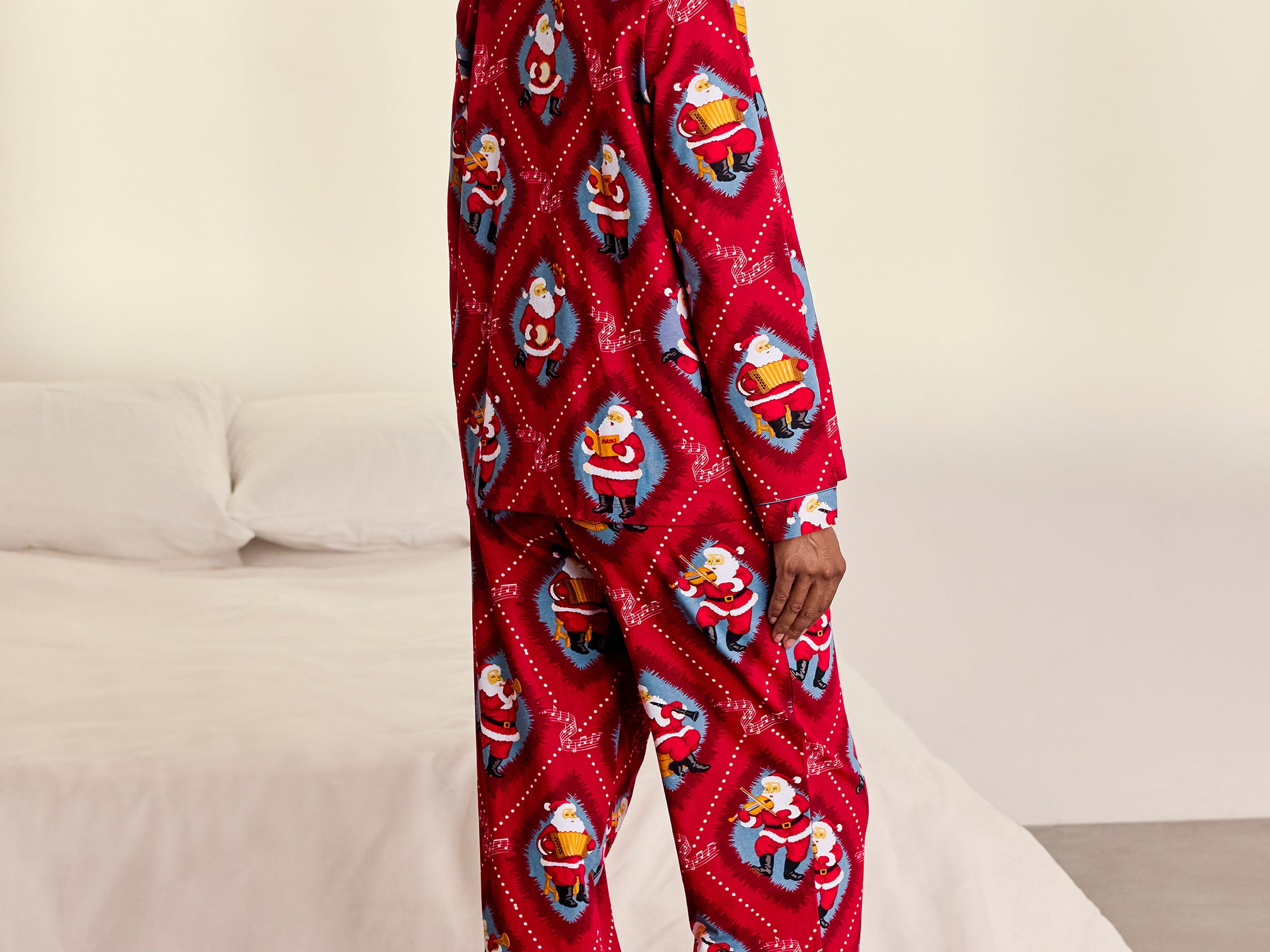 A woman stands barefoot in a bedroom wearing a red long-sleeve pajama one-piece covered in a repeating Santa Claus print. A white bed and light wall are visible in the background.