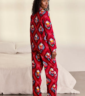 A woman stands barefoot in a bedroom wearing a red long-sleeve pajama one-piece covered in a repeating Santa Claus print. A white bed and light wall are visible in the background.