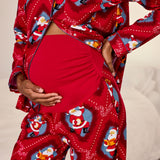 A close-up of a pregnant person wearing a red pajama set with a festive Santa Claus print. The long-sleeve top is buttoned and open, showing the belly, while the matching pants and a solid red belly band are also visible against a soft neutral background.