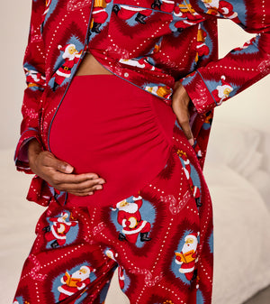 A close-up of a pregnant person wearing a red pajama set with a festive Santa Claus print. The long-sleeve top is buttoned and open, showing the belly, while the matching pants and a solid red belly band are also visible against a soft neutral background.
