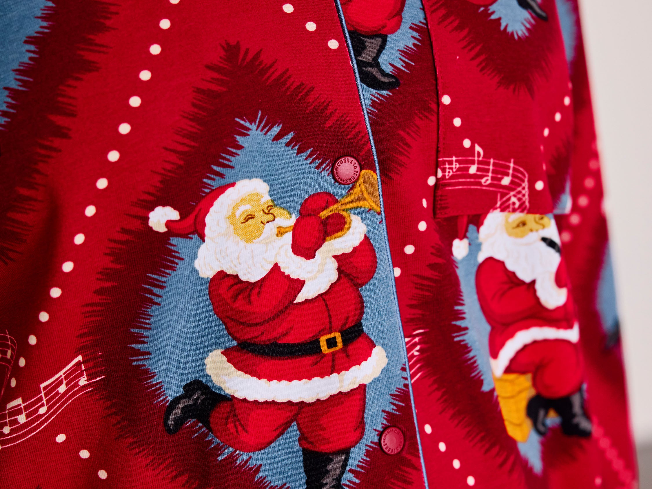 A close-up of a red Christmas-themed button-up shirt featuring a repeating print of Santa Claus playing a trumpet inside blue circular frames, with white musical notes and dots on a red background.