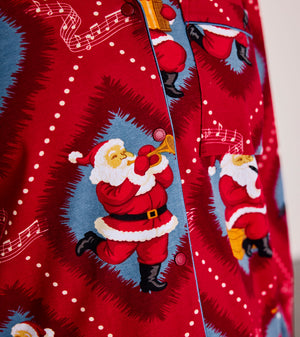 A close-up of a red Christmas-themed button-up shirt featuring a repeating print of Santa Claus playing a trumpet inside blue circular frames, with white musical notes and dots on a red background.