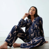 A woman sits barefoot on a light floor wearing a navy blue pajama set with a beige giraffe print. The set includes a long-sleeve button-up shirt and matching pants.