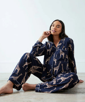 A woman sits barefoot on a light floor wearing a navy blue pajama set with a beige giraffe print. The set includes a long-sleeve button-up shirt and matching pants.