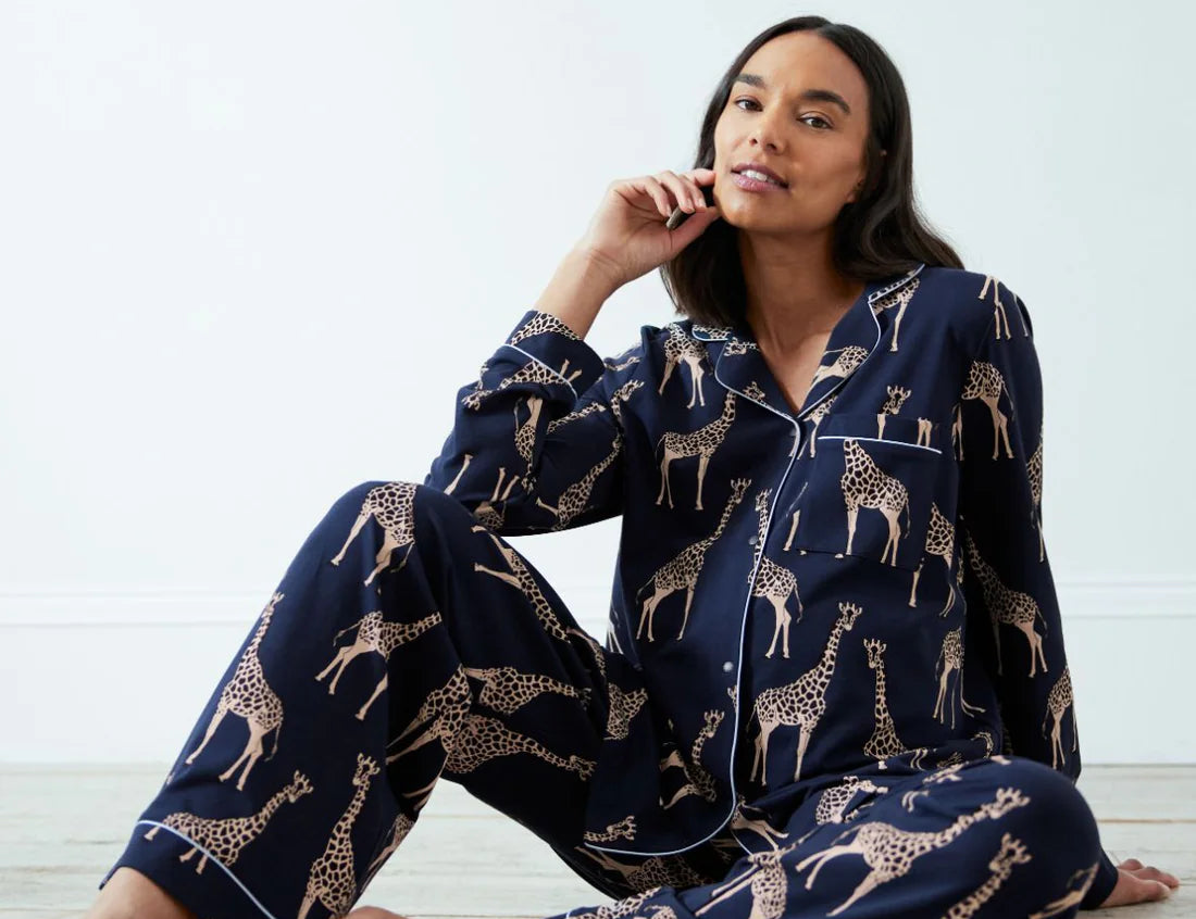 A woman sits barefoot on a light floor wearing a navy blue pajama set with a beige giraffe print. The set includes a long-sleeve button-up shirt and matching pants.