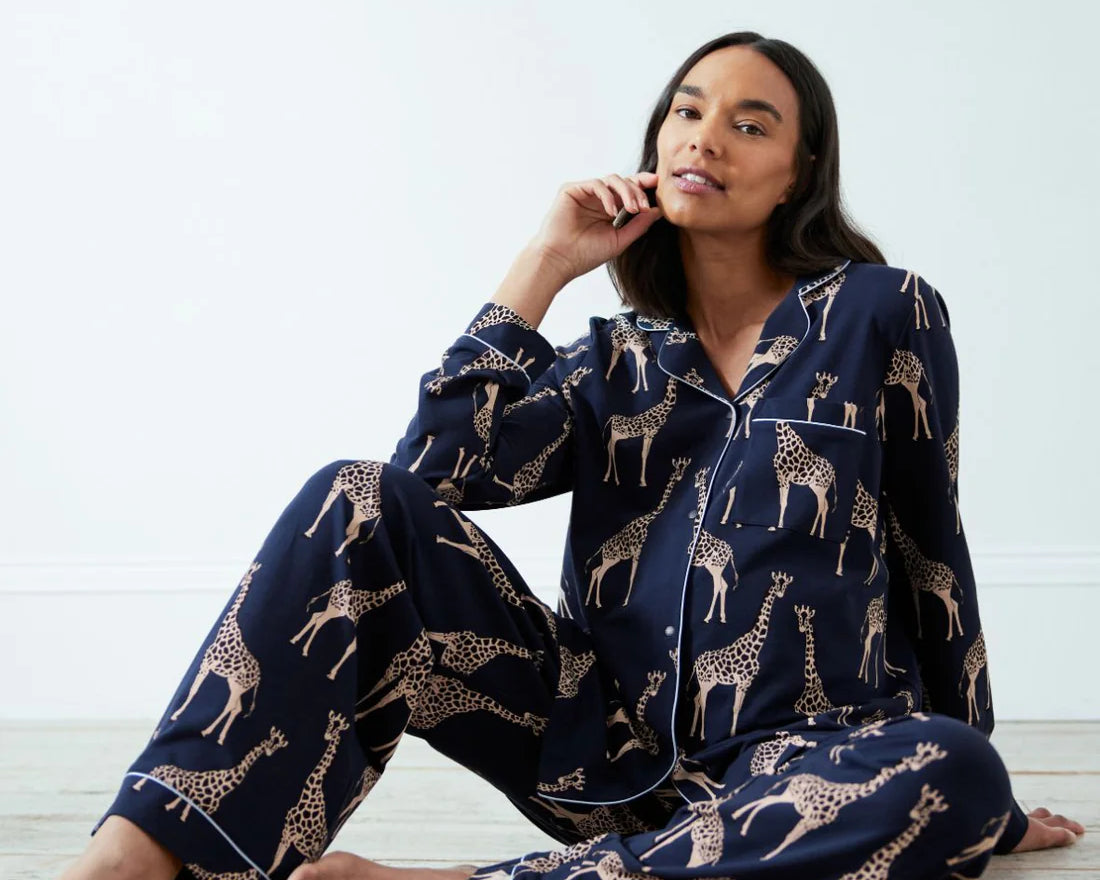 A woman sits barefoot on a light floor wearing a navy blue pajama set with a beige giraffe print. The set includes a long-sleeve button-up shirt and matching pants.