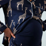 A close-up of a person wearing navy blue pajama set with a beige giraffe print and light piping. The image shows the torso, hands, and a pregnant belly wearing the matching top and skirt.
