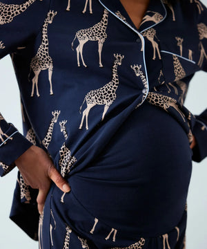 A close-up of a person wearing navy blue pajama set with a beige giraffe print and light piping. The image shows the torso, hands, and a pregnant belly wearing the matching top and skirt.