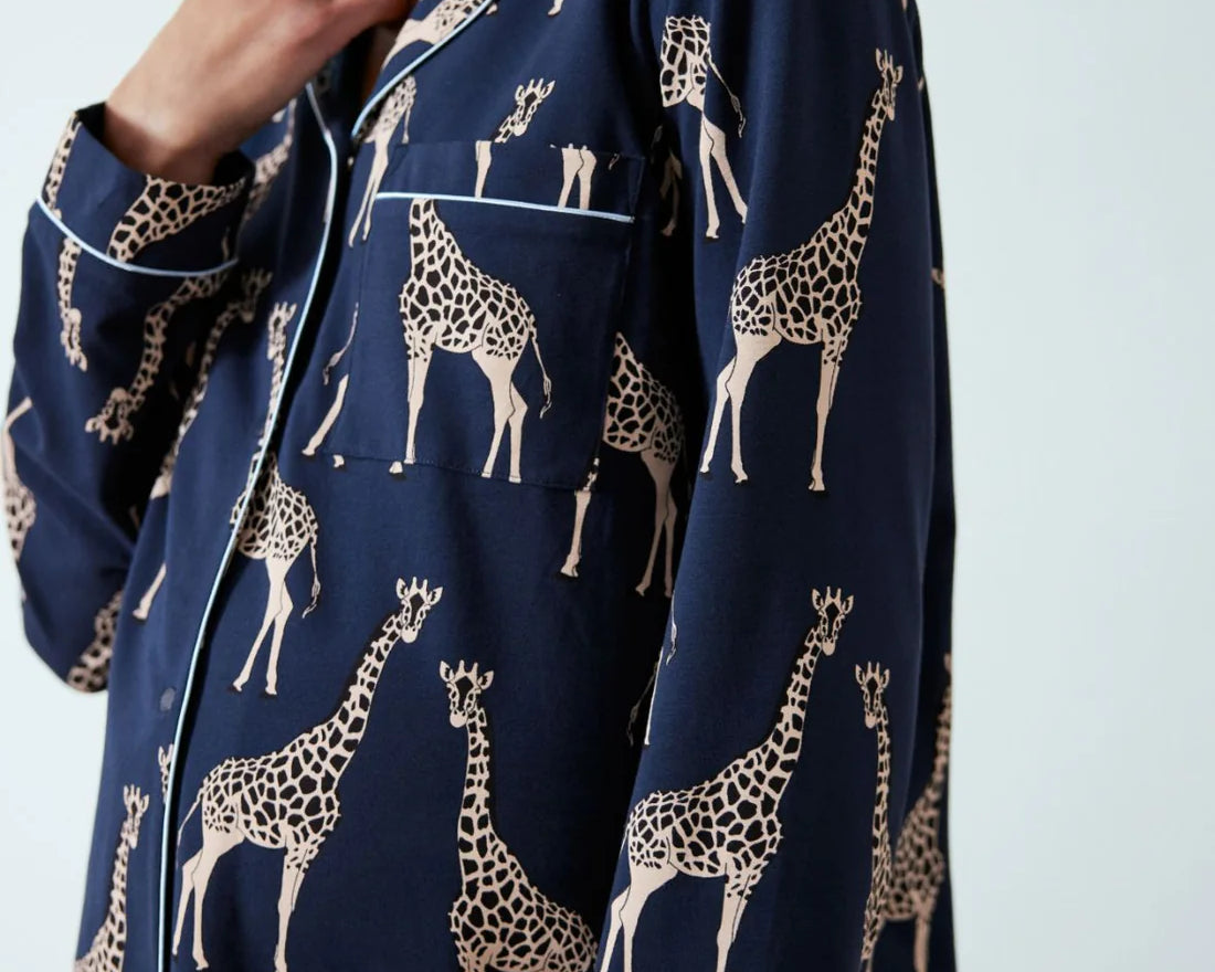 A close-up of a person wearing a navy blue pajama shirt with an all-over beige giraffe print and white piping details.