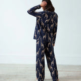 A woman stands with her back to the camera wearing a navy blue pajama set with a beige giraffe print, set against a plain white wall and light wood floor.