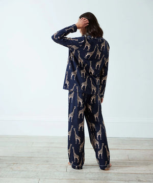 A woman stands with her back to the camera wearing a navy blue pajama set with a beige giraffe print, set against a plain white wall and light wood floor.