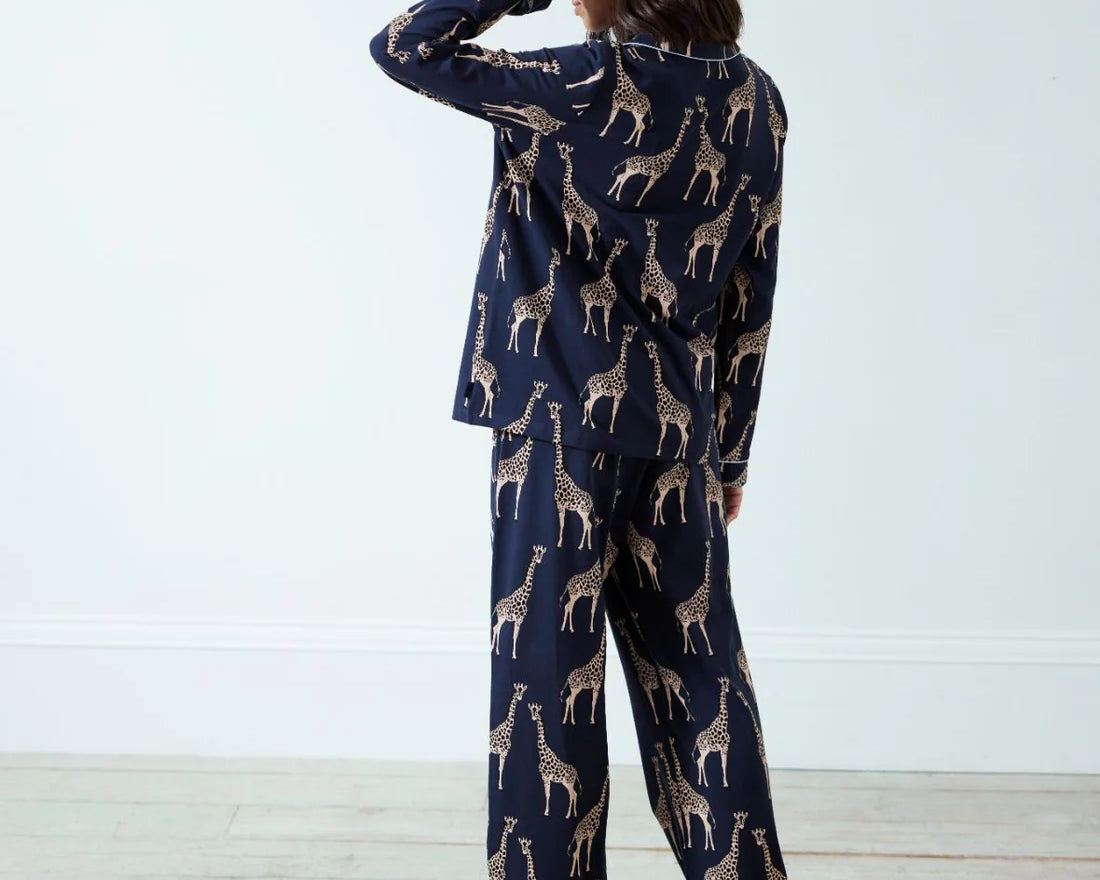 A woman stands with her back to the camera wearing a navy blue pajama set with a beige giraffe print, set against a plain white wall and light wood floor.