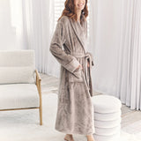 A woman wearing a plush, long taupe-gray bathrobe stands barefoot in a bright, modern bedroom setting with white curtains, a textured chair, and a round ottoman.