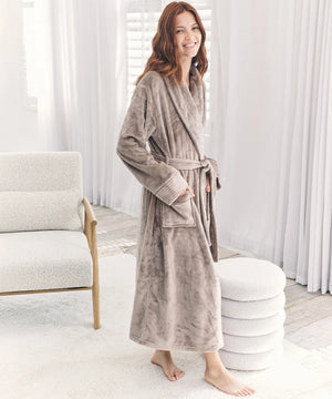 A woman wearing a plush, long taupe-gray bathrobe stands barefoot in a bright, modern bedroom setting with white curtains, a textured chair, and a round ottoman.