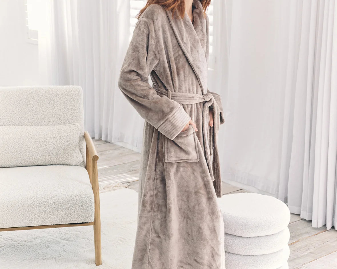A woman wearing a plush, long taupe-gray bathrobe stands barefoot in a bright, modern bedroom setting with white curtains, a textured chair, and a round ottoman.