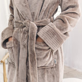 A close-up of a person wearing a plush bathrobe in a taupe-brown color. The robe features a tie belt, ribbed cuffs, and front patch pockets with the person's hands inside.