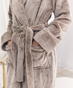 A close-up of a person wearing a plush bathrobe in a taupe-brown color. The robe features a tie belt, ribbed cuffs, and front patch pockets with the person's hands inside.