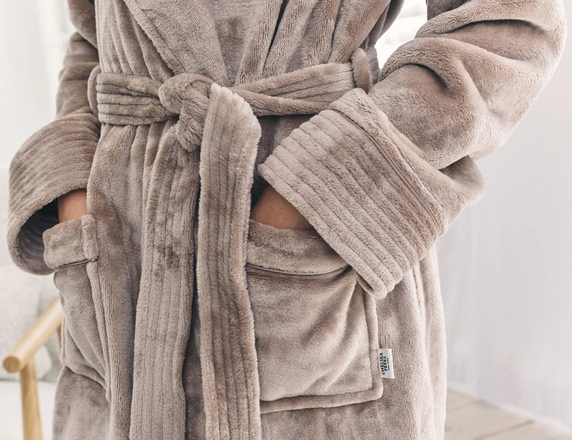 A close-up of a person wearing a plush bathrobe in a taupe-brown color. The robe features a tie belt, ribbed cuffs, and front patch pockets with the person's hands inside.