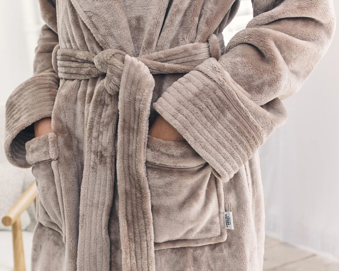 A close-up of a person wearing a plush bathrobe in a taupe-brown color. The robe features a tie belt, ribbed cuffs, and front patch pockets with the person's hands inside.
