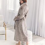 A woman wearing a plush, long taupe-gray bathrobe stands barefoot in a bright, modern room with white curtains and a light-colored chair.