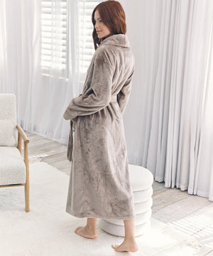 A woman wearing a plush, long taupe-gray bathrobe stands barefoot in a bright, modern room with white curtains and a light-colored chair.