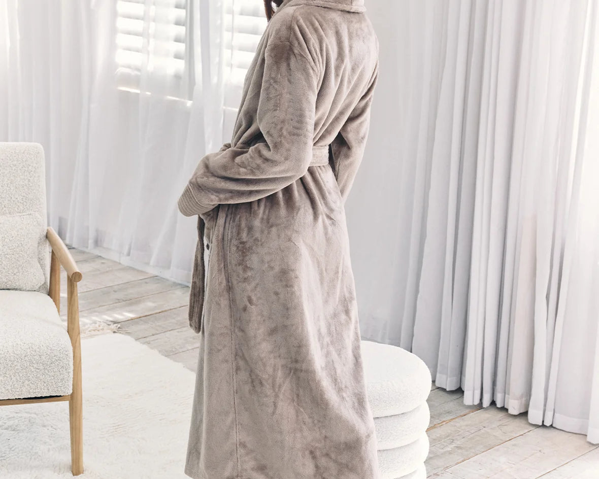 A woman wearing a plush, long taupe-gray bathrobe stands barefoot in a bright, modern room with white curtains and a light-colored chair.