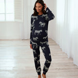 A woman modeling a navy blue long-sleeve pajama set with a white zebra print, standing in a bright room with white curtains and light wood floors.
