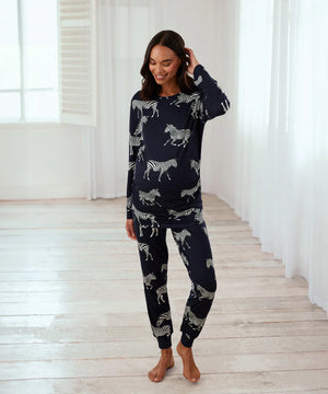 A woman modeling a navy blue long-sleeve pajama set with a white zebra print, standing in a bright room with white curtains and light wood floors.