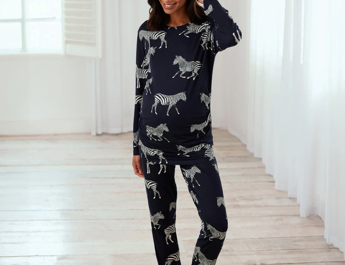 A woman modeling a navy blue long-sleeve pajama set with a white zebra print, standing in a bright room with white curtains and light wood floors.