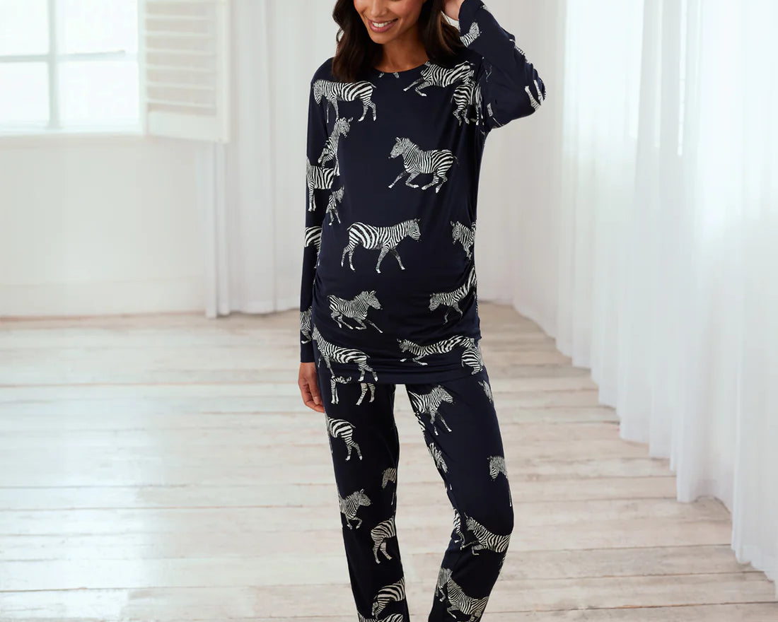 A woman modeling a navy blue long-sleeve pajama set with a white zebra print, standing in a bright room with white curtains and light wood floors.