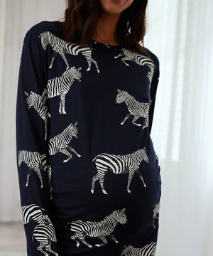 A woman wearing a navy blue long-sleeve top with an all-over white zebra print, shown from the waist up against a bright white curtain background.