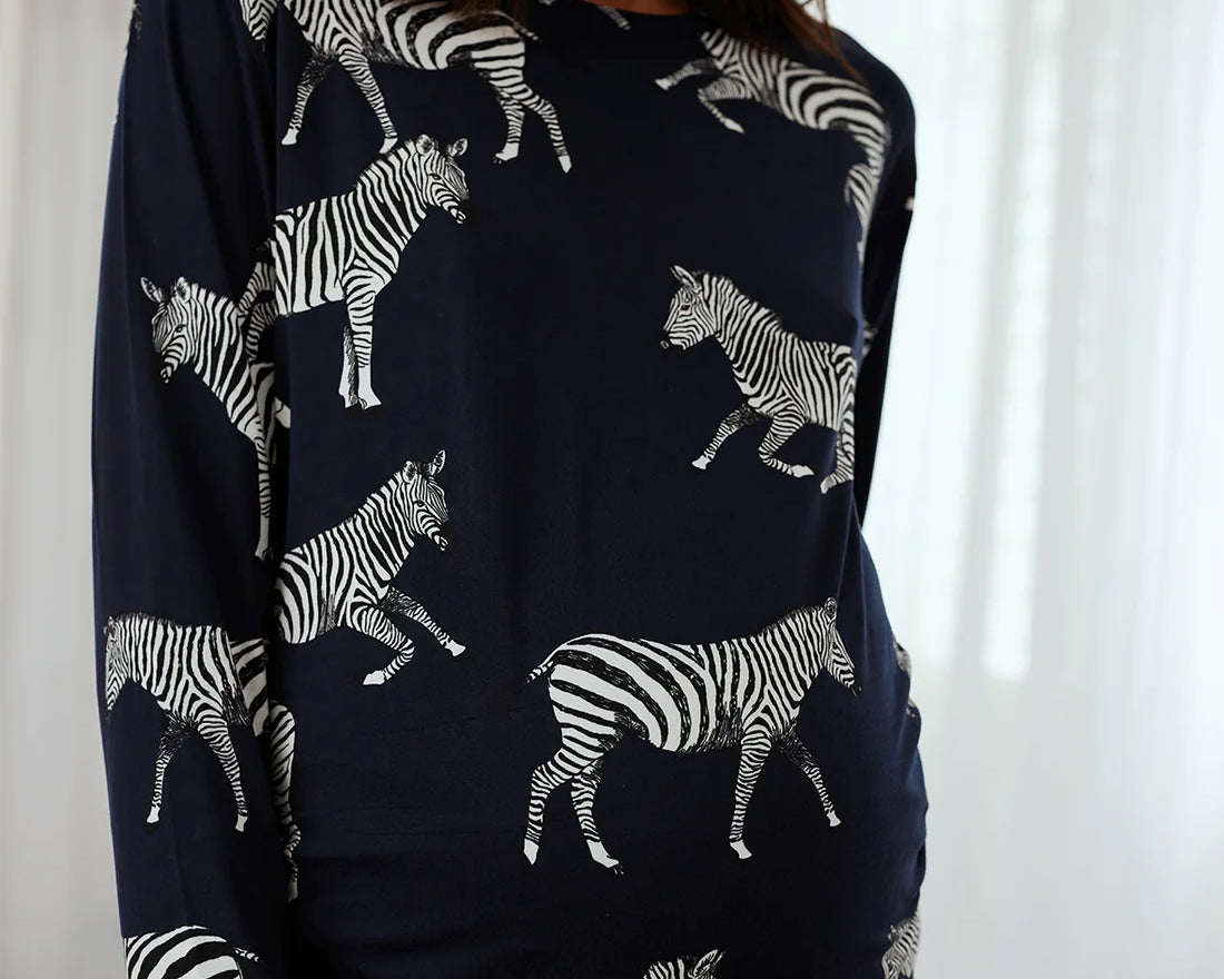 A woman wearing a navy blue long-sleeve top with an all-over white zebra print, shown from the waist up against a bright white curtain background.
