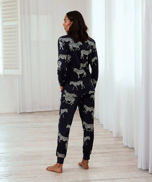 A woman wearing a matching two-piece pajama set with a zebra print, standing barefoot in a bright room with white curtains and light wood floors.