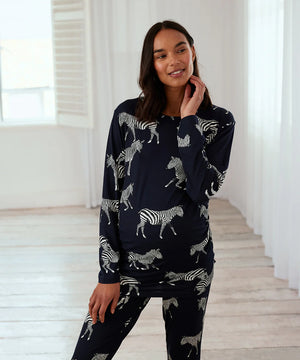 A woman modeling a matching two-piece pajama set featuring a dark navy blue base with a white zebra print, posed in a bright, minimalist room with white walls and light wood flooring.